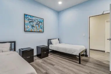 A shared patient room at Boca Recovery Center Huntington, with two beds, nightstands, and a colorful world map artwork on the wall. The room has soft blue walls, wood-like flooring, and a door leading to another area.