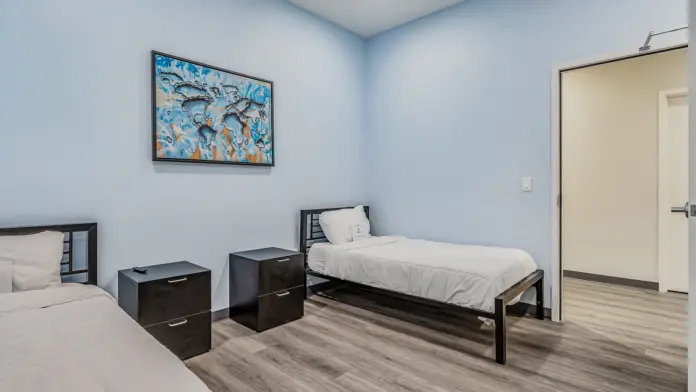 A shared patient room at Boca Recovery Center Huntington, with two beds, nightstands, and a colorful world map artwork on the wall. The room has soft blue walls, wood-like flooring, and a door leading to another area.