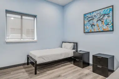 A simple patient room at Boca Recovery Center Huntington, featuring a bed, two nightstands, and a colorful world map artwork on the wall. The room has soft blue walls and a window with blinds.