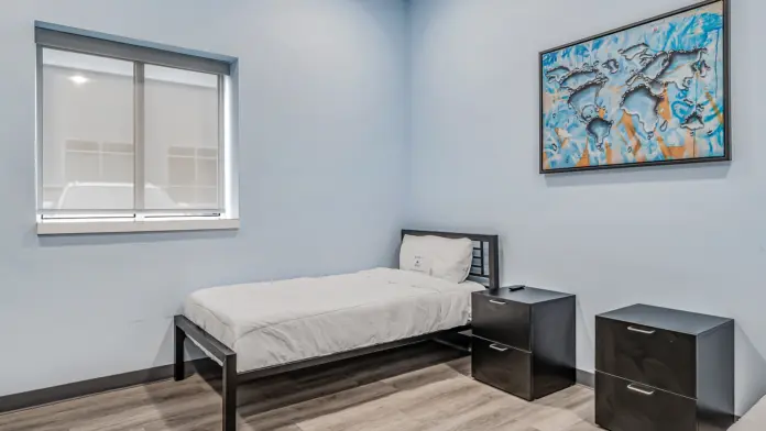 A simple patient room at Boca Recovery Center Huntington, featuring a bed, two nightstands, and a colorful world map artwork on the wall. The room has soft blue walls and a window with blinds.