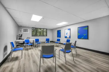 A bright, modern group therapy room with a circle of blue chairs, wood flooring, a computer desk in the corner, and abstract artwork on the walls.