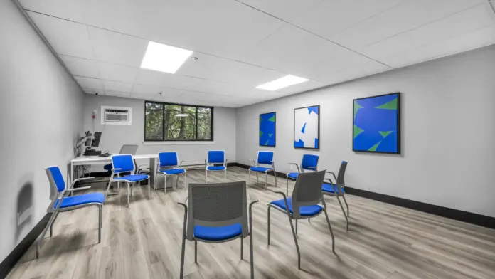 A bright, modern group therapy room with a circle of blue chairs, wood flooring, a computer desk in the corner, and abstract artwork on the walls.