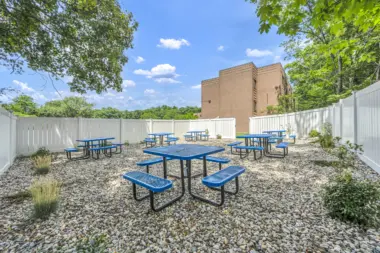 An outdoor recreation area with blue metal picnic tables on a gravel surface, surrounded by white privacy fencing and greenery, under a clear blue sky.