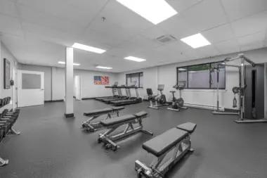 A clean, modern fitness center with weight benches, dumbbells, treadmills, stationary bikes, and a cable machine, featuring bright lighting and an American flag wall art.