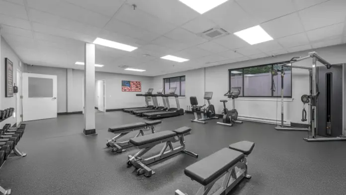 A clean, modern fitness center with weight benches, dumbbells, treadmills, stationary bikes, and a cable machine, featuring bright lighting and an American flag wall art.