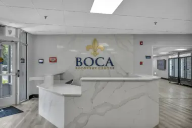 A sleek and modern reception area at Boca Recovery Center featuring a marble-style front desk and wall with the center’s logo, located near a glass entrance and adjacent cafeteria.