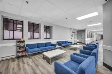 A modern lounge area with blue sofas and armchairs, white coffee tables, and a rack of books and games, located next to a hallway lined with framed artwork