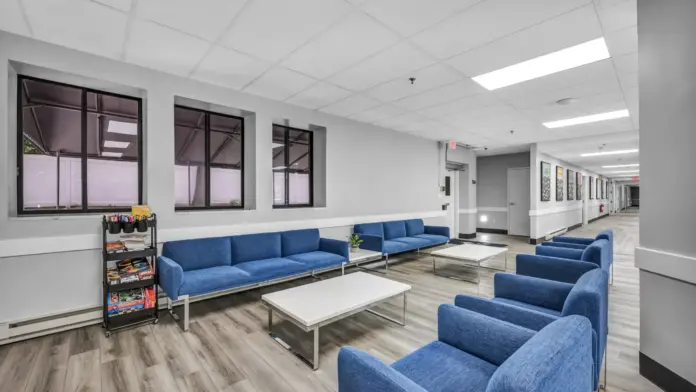A modern lounge area with blue sofas and armchairs, white coffee tables, and a rack of books and games, located next to a hallway lined with framed artwork