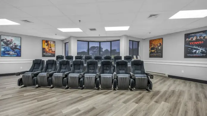 A private movie theater room with two rows of plush reclining chairs, wood flooring, and movie posters on the walls, creating a cozy entertainment space.