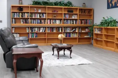 Reading room or library with large wooden bookshelves filled with books. A gray leather recliner and a small coffee table with a globe sit in front of the shelves. A smaller bookshelf is on the right, and a world map hangs on the wall. The room has a gray and white tiled floor.