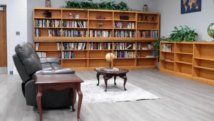 Reading room or library with large wooden bookshelves filled with books. A gray leather recliner and a small coffee table with a globe sit in front of the shelves. A smaller bookshelf is on the right, and a world map hangs on the wall. The room has a gray and white tiled floor.