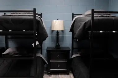 Dorm room interior with two bunk beds, each with dark gray bedding and a gray pillow. A black nightstand with two drawers and a lamp sits between the beds. The walls are light blue cinder block.