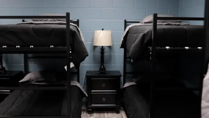 Dorm room interior with two bunk beds, each with dark gray bedding and a gray pillow. A black nightstand with two drawers and a lamp sits between the beds. The walls are light blue cinder block.