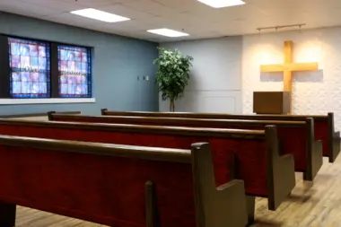 Interior view of a small chapel or meditation room with rows of empty wooden pews facing a textured wall with a brightly lit wooden cross. Stained-glass windows with the words 