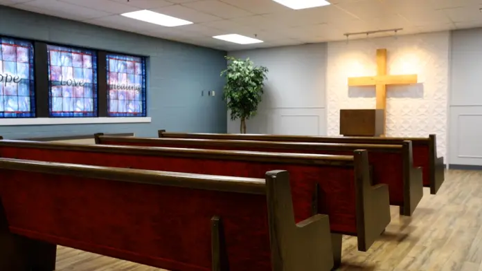 Interior view of a small chapel or meditation room with rows of empty wooden pews facing a textured wall with a brightly lit wooden cross. Stained-glass windows with the words