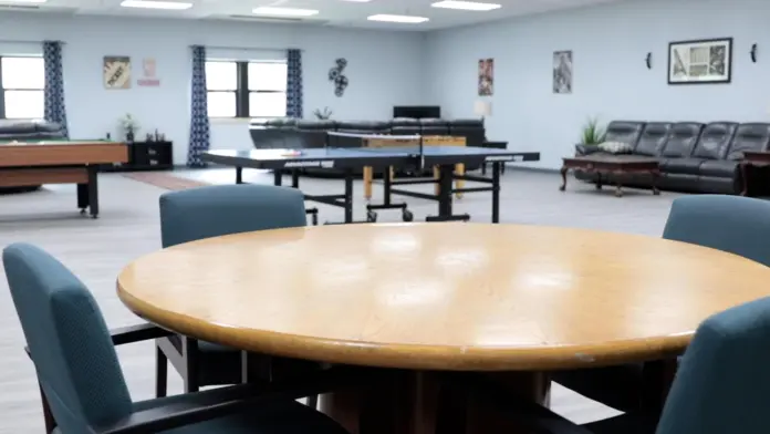 Large recreation room with light blue walls, featuring a billiards table on the left, two ping pong tables in the center, and a seating area with dark leather sofas on the right. A round wooden table with four blue chairs is in the foreground. The room has a light-colored floor and recessed lighting. Artwork is visible on the walls.
