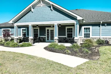 Exterior view of a two-story building with blue siding and a prominent front porch. The porch features white columns and a set of double doors at the center. A paved walkway leads to the entrance, and landscaping includes bushes and patches of lawn. The sky is clear and blue.