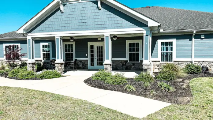 Exterior view of a two-story building with blue siding and a prominent front porch. The porch features white columns and a set of double doors at the center. A paved walkway leads to the entrance, and landscaping includes bushes and patches of lawn. The sky is clear and blue.