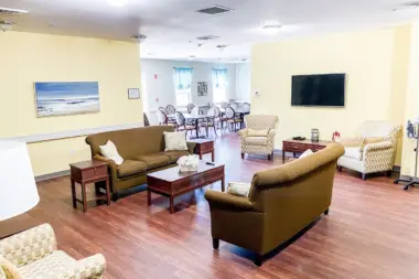 Spacious common room with yellow walls, hardwood floors, and comfortable seating. Two brown sofas face each other, flanked by patterned armchairs. A coffee table sits in the center, and a large television is mounted on the wall. A dining area with a round table and chairs is visible in the background through an open doorway.