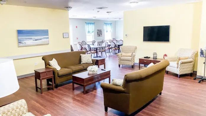 Spacious common room with yellow walls, hardwood floors, and comfortable seating. Two brown sofas face each other, flanked by patterned armchairs. A coffee table sits in the center, and a large television is mounted on the wall. A dining area with a round table and chairs is visible in the background through an open doorway.