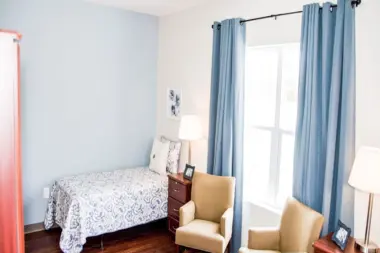 Serene bedroom interior with light blue walls, two armchairs, and a single bed with patterned bedding. A window with blue curtains overlooks a bright outdoor scene. Two side tables with lamps flank the bed, and a wardrobe is visible on the left.