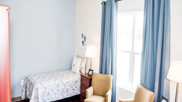 Serene bedroom interior with light blue walls, two armchairs, and a single bed with patterned bedding. A window with blue curtains overlooks a bright outdoor scene. Two side tables with lamps flank the bed, and a wardrobe is visible on the left.