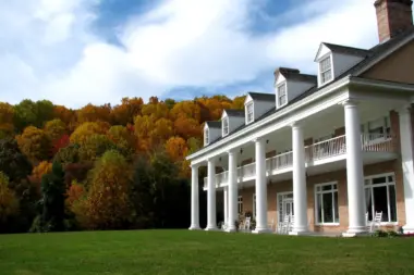 Large, elegant building with white columns and a red brick facade, set against a backdrop of vibrant autumn foliage. The building features a multi-tiered portico with white columns and railings, and dormer windows in the pitched roof. A well-maintained green lawn stretches out in front, and a single chimney rises from the right side of the building. The sky is partly cloudy with patches of blue.