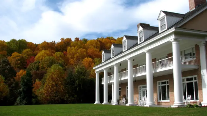 Large, elegant building with white columns and a red brick facade, set against a backdrop of vibrant autumn foliage. The building features a multi-tiered portico with white columns and railings, and dormer windows in the pitched roof. A well-maintained green lawn stretches out in front, and a single chimney rises from the right side of the building. The sky is partly cloudy with patches of blue.