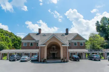 Large, stately brick building with a central arched entrance and a gray shingled roof. The building features multiple chimneys, evenly spaced windows, and a covered walkway extending along the front. Several cars are parked in the paved area in front of the building. The sky is bright blue with fluffy white clouds, and trees are visible in the background.