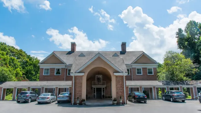 Large, stately brick building with a central arched entrance and a gray shingled roof. The building features multiple chimneys, evenly spaced windows, and a covered walkway extending along the front. Several cars are parked in the paved area in front of the building. The sky is bright blue with fluffy white clouds, and trees are visible in the background.