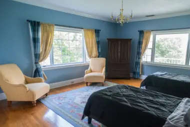 Bedroom with blue walls, featuring two twin beds with dark comforters, two armchairs, and a large wooden wardrobe. Two windows with blue and gold curtains flank a central armchair. A smaller armchair sits to the left. A light blue and pink rug covers part of the hardwood floor. A chandelier hangs from the white ceiling.
