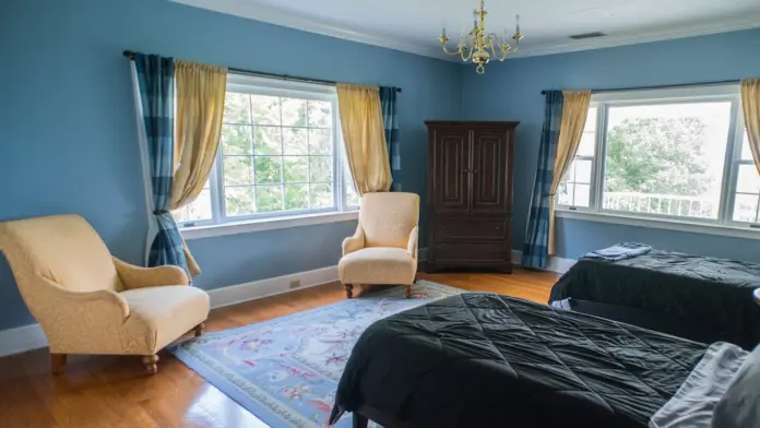 Bedroom with blue walls, featuring two twin beds with dark comforters, two armchairs, and a large wooden wardrobe. Two windows with blue and gold curtains flank a central armchair. A smaller armchair sits to the left. A light blue and pink rug covers part of the hardwood floor. A chandelier hangs from the white ceiling.