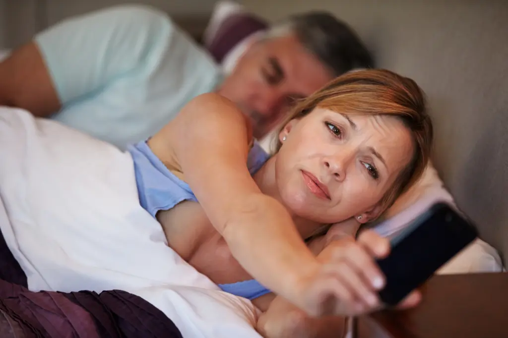 A woman stares at her phone in the middle of the night, having trouble sleeping or suffering from insomnia