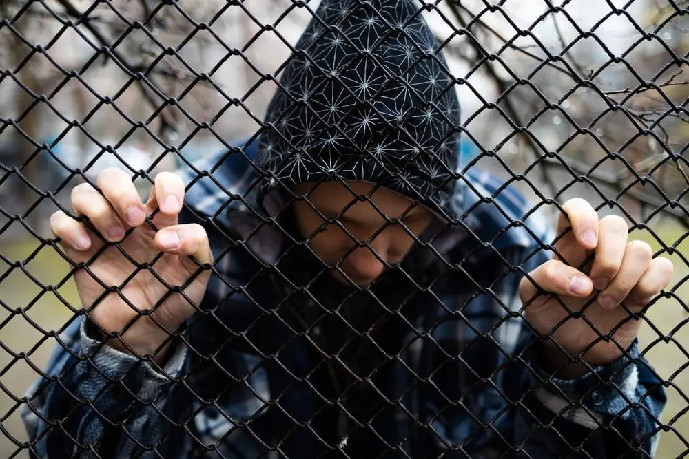 A depressed man in a hoodie grips a chainlink fence. Learn more about xylazine (tranq) addiction here