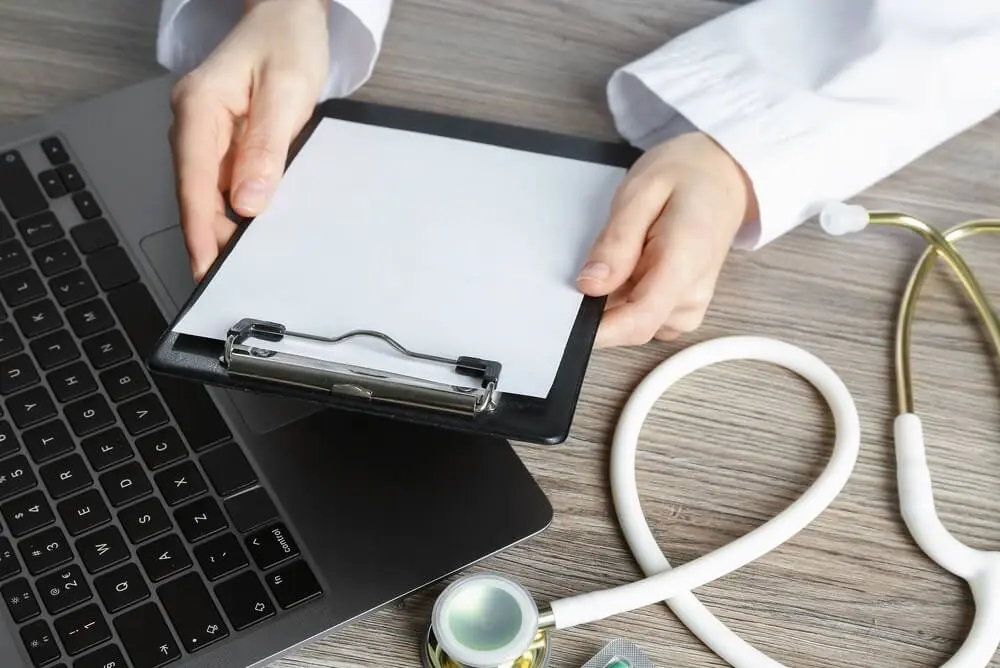 A doctor holding a clipboard next to a laptop. Learn more about virtual and online IOP drug rehab, here.