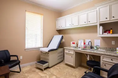 Interior examination room with medical chair, white cabinetry, countertop supplies, and window.