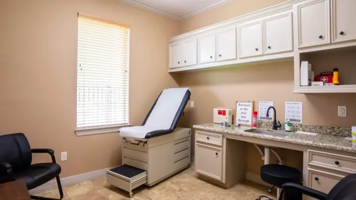 Interior examination room with medical chair, white cabinetry, countertop supplies, and window.