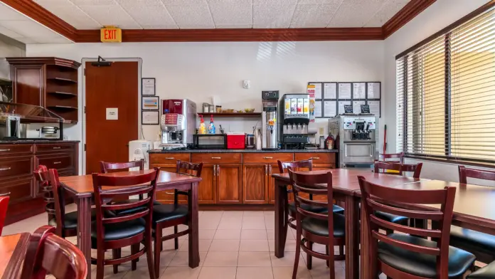 Interior dining space with wooden tables and chairs, coffee station, appliances, and large window with blinds.