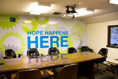 Interior meeting room with conference table, office chairs, ceiling projector, and green gear mural with hope message
