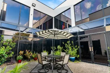 Exterior patio area with striped umbrellas, dining table with chairs, landscaping, and facility building visible in background