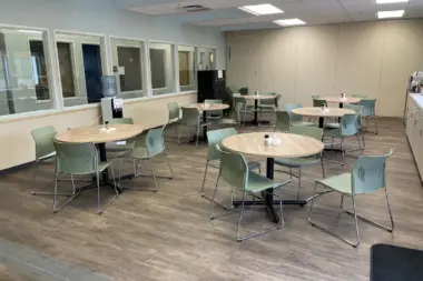 Interior multipurpose room with round tables, green upholstered chairs, and glass-walled office areas along perimeter.