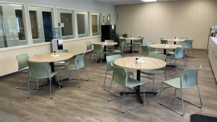 Interior multipurpose room with round tables, green upholstered chairs, and glass-walled office areas along perimeter.
