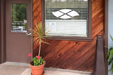 Exterior entrance detail showing brown door with posted dosing hours, window with blinds, and orange flowering plants