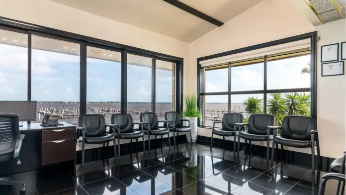 Interior high-rise office with floor-to-ceiling windows, black office chairs, conference tables, and panoramic waterfront cityscape view.