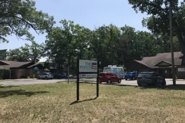 Outdoor facility sign mounted on post with campus buildings, parked vehicles, and trees in background