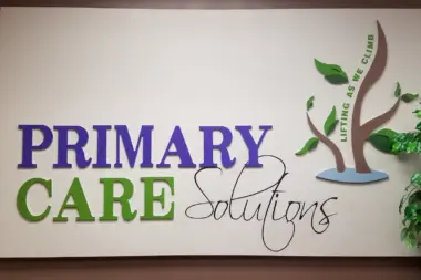 Interior signage showing Primary Care Solutions logo with green and blue tree graphic and tagline on white wall