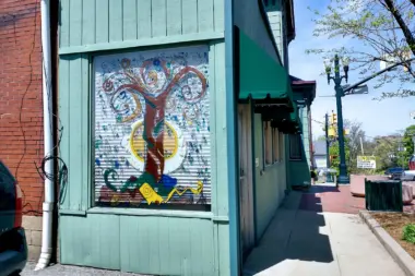 Exterior teal building with colorful tree mural artwork, brick adjoining structure, sidewalk frontage with street and parked vehicles visible.