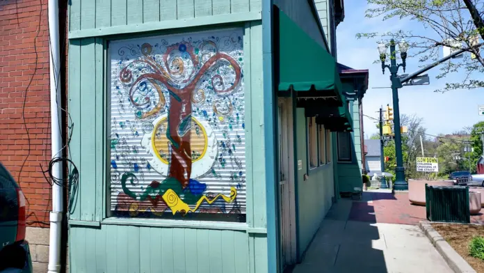 Exterior teal building with colorful tree mural artwork, brick adjoining structure, sidewalk frontage with street and parked vehicles visible.