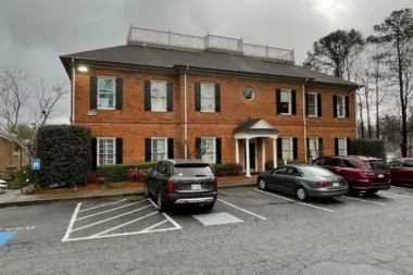 Two-story brick building with white column entry and manicured landscaping, visitor parking area visible