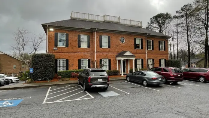 Two-story brick building with white column entry and manicured landscaping, visitor parking area visible